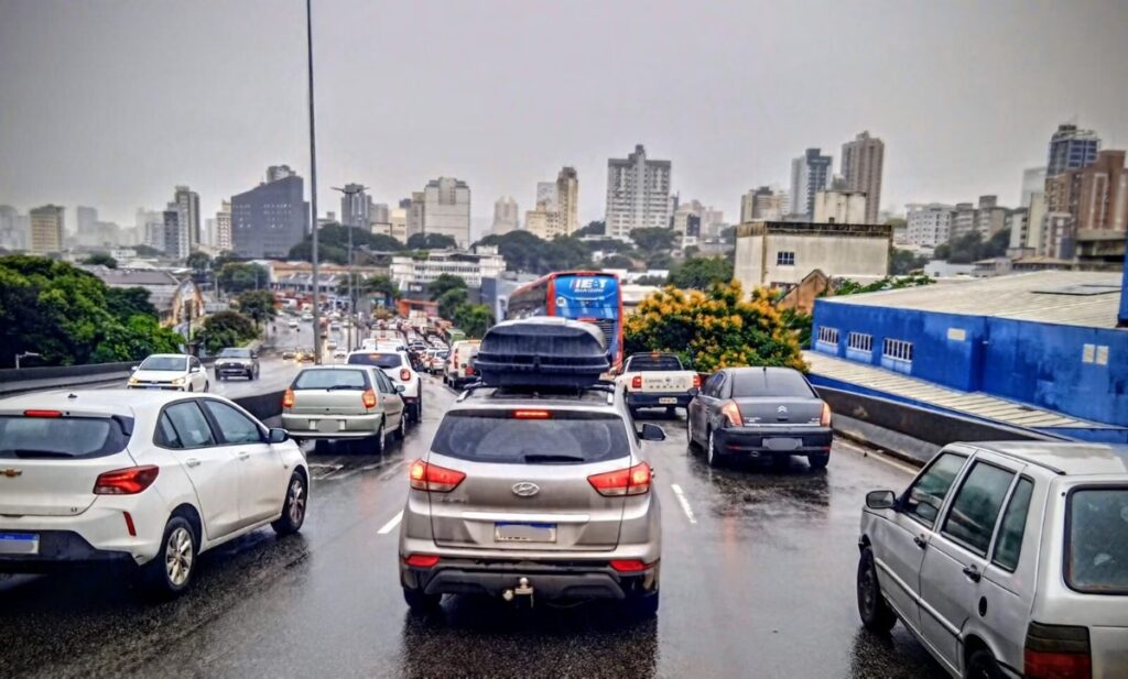 Fila de carros e ônibus em via urbana sob chuva, com asfalto molhado e prédios ao fundo.