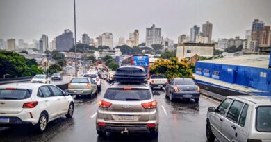 Fila de carros e ônibus em via urbana sob chuva, com asfalto molhado e prédios ao fundo.
