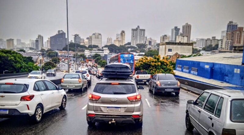 Fila de carros e ônibus em via urbana sob chuva, com asfalto molhado e prédios ao fundo.